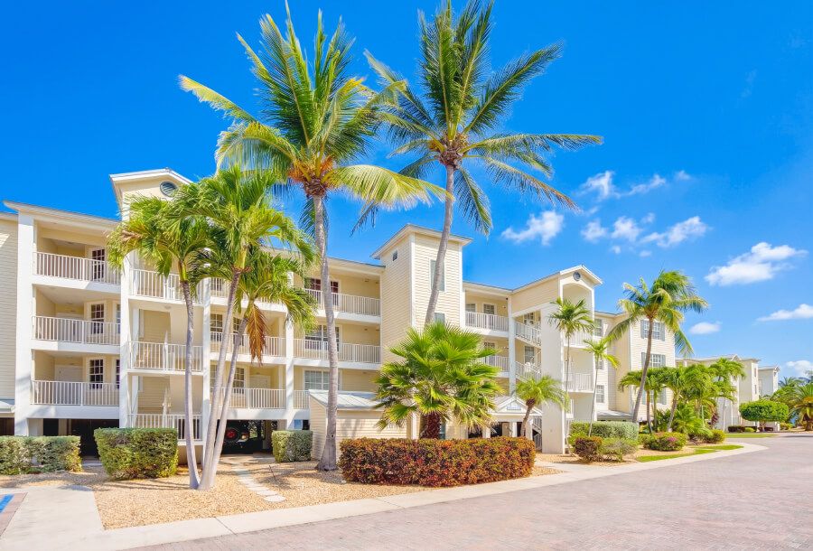 Beige, three-story apartment building with white railings and balconies, fronted by palm trees and a sunny blue sky.